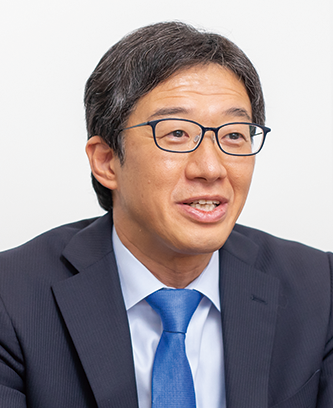 A professional portrait of a man in a suit and glasses, associated with Tokio Marine Nichido. He is wearing a blue tie and speaking, set against a plain background.