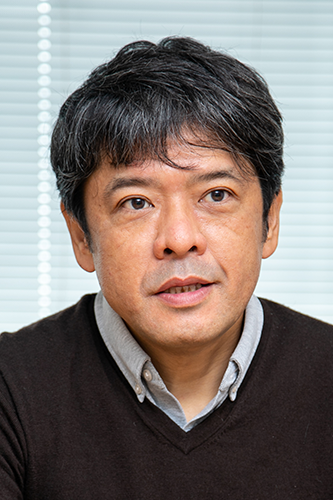 Portrait of a man with dark hair wearing a brown sweater and light collared shirt, sitting in front of a background with horizontal blinds.